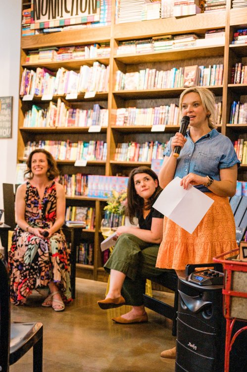 A woman speaks into a microphone while two others listen in a cozy bookstore filled with colorful books.