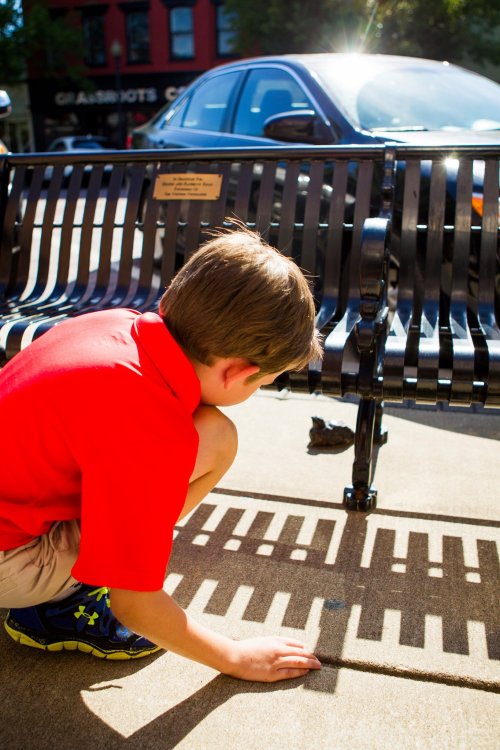 A boy in a red shirt crouches by a bench, observing shadows on the ground in bright sunlight.