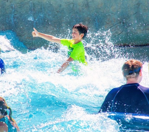 A boy in a bright green shirt gives a thumbs up while splashing in a pool with other children.