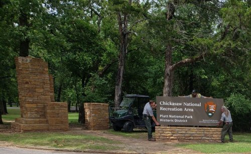 Two park rangers work on a stone sign for Chickasaw National Recreation Area, surrounded by trees and greenery.