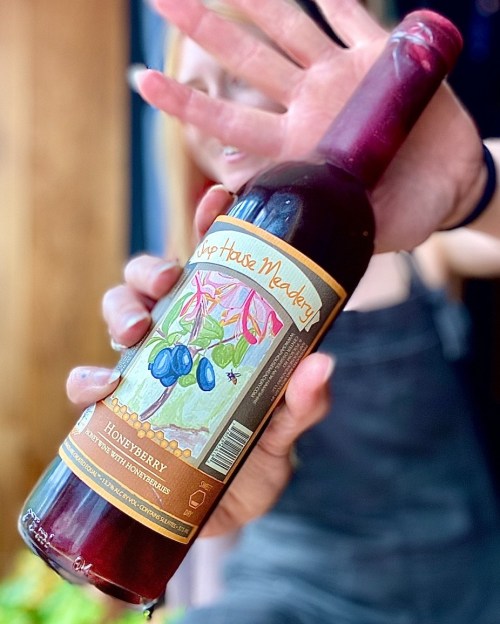 A person holds a bottle of Honeyberry mead with a colorful label featuring flowers and berries.