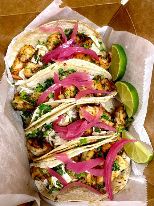 A takeout box filled with shrimp tacos topped with pickled onions, cilantro, and lime wedges.