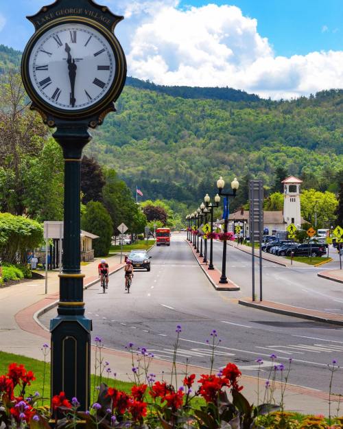 A scenic street view with a clock, cyclists, flowers, and mountains under a blue sky with fluffy clouds.