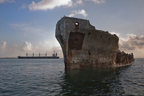 A rusted shipwreck in calm waters, with a cargo ship in the background under a cloudy sky.