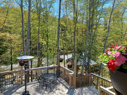 A wooden deck surrounded by trees, featuring a table and chairs, with a gazebo visible in the background.