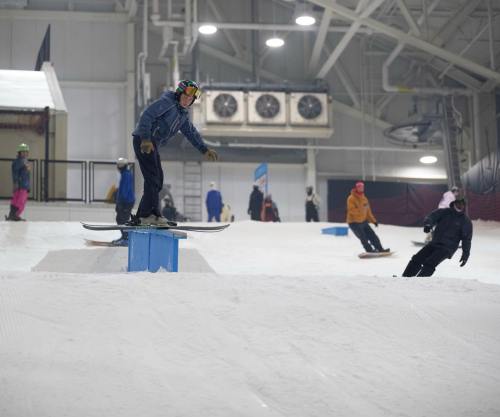 A snowboarder performs a trick on a rail in an indoor snow park, with others riding in the background.