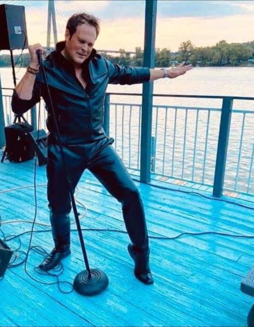 A performer in a black jumpsuit poses dramatically with a microphone on a waterfront stage at sunset.