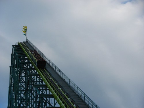 A roller coaster climbs steeply towards the sky, with flags waving at the top against a cloudy backdrop.
