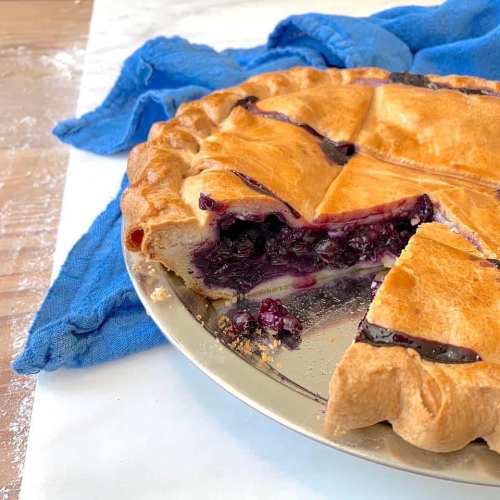 A freshly baked blueberry pie on a silver plate, with a slice removed, resting on a blue cloth.