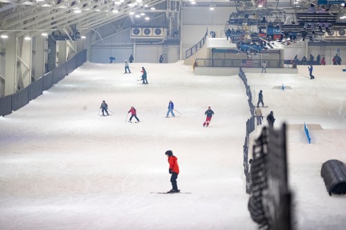 Indoor ski slope with people skiing and snowboarding on a snowy surface, surrounded by safety barriers.