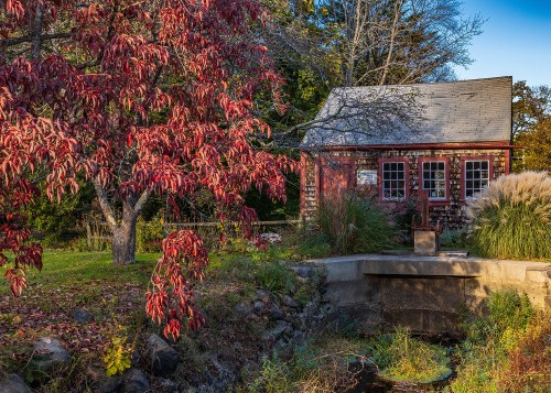 A quaint red house surrounded by vibrant autumn foliage and a small stream, creating a serene countryside scene.