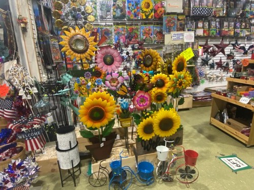 A vibrant display of colorful metal flowers and garden decorations in a shop, with various crafts and items in the background.