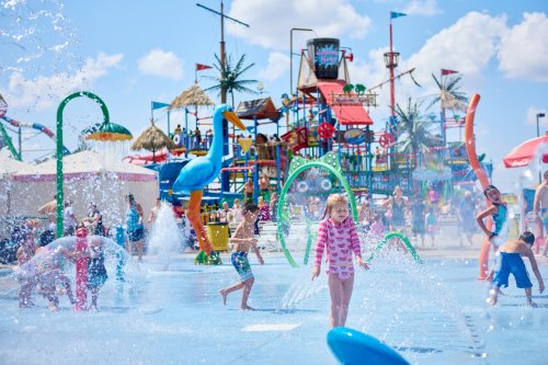 A vibrant water park scene with children playing in fountains, colorful slides, and tropical decorations under a sunny sky.
