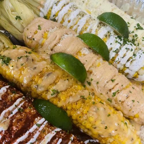 A close-up of elote, grilled corn on the cob topped with sauces, cheese, and lime wedges, garnished with parsley.