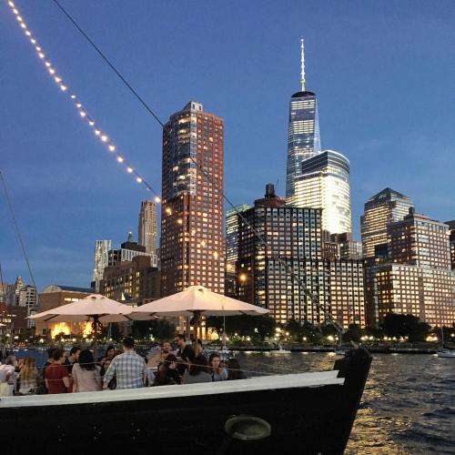 A lively waterfront scene with people dining outdoors, illuminated city skyline, and a tall skyscraper at dusk.