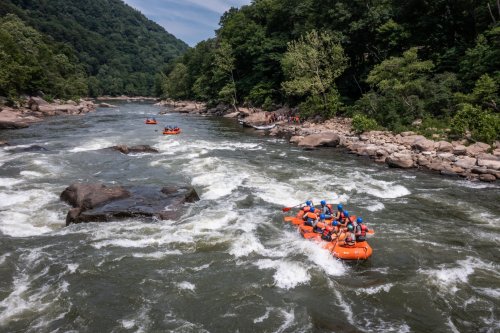 A group of people in orange rafts navigating a river with rocky banks and lush green trees in the background.