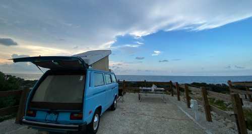 A blue camper van parked near the ocean under a cloudy sky, with a picnic table in the foreground.