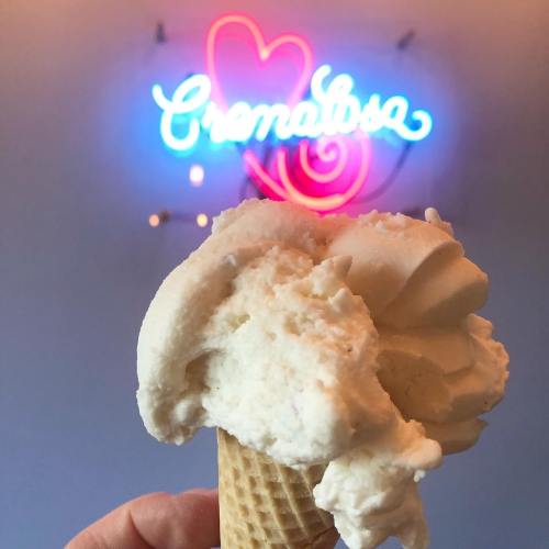 A hand holding a cone of creamy ice cream in front of a colorful neon sign that reads "Cremalosa."