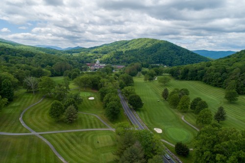 Aerial view of a lush green golf course surrounded by rolling hills and trees under a cloudy sky.