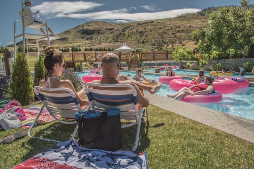 Two people relax on lounge chairs by a pool, surrounded by others enjoying inflatable floats and a sunny landscape.