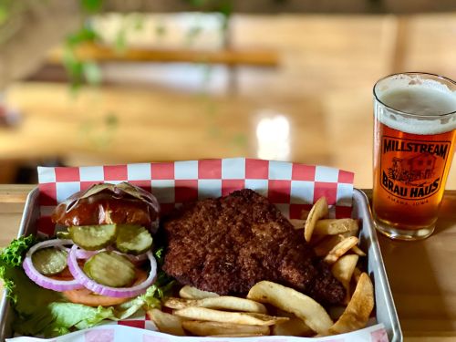 A plate with a crispy fried chicken sandwich, pickles, lettuce, fries, and a glass of beer on a wooden table.