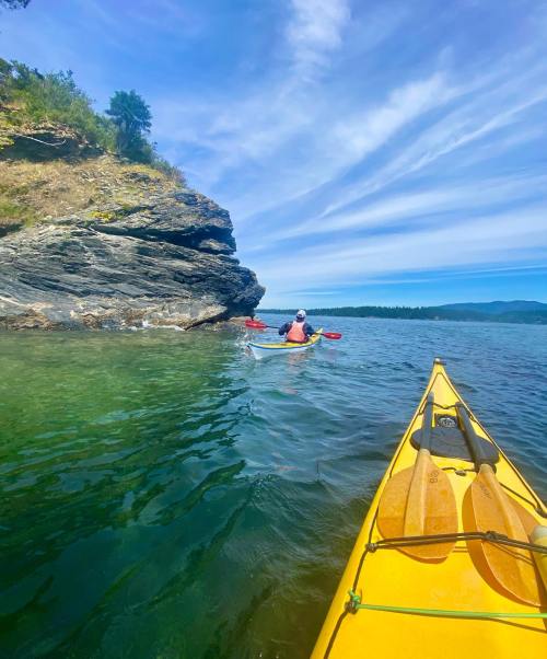 A yellow kayak in the foreground with a person paddling in the background near rocky cliffs and clear water.