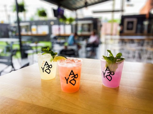 Three colorful cocktails on a wooden table, featuring lime and mint garnishes, in a vibrant outdoor setting.