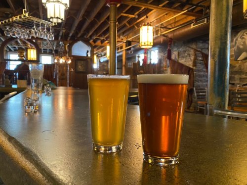 Two glasses of beer, one golden and one amber, sit on a bar counter in a rustic pub setting.