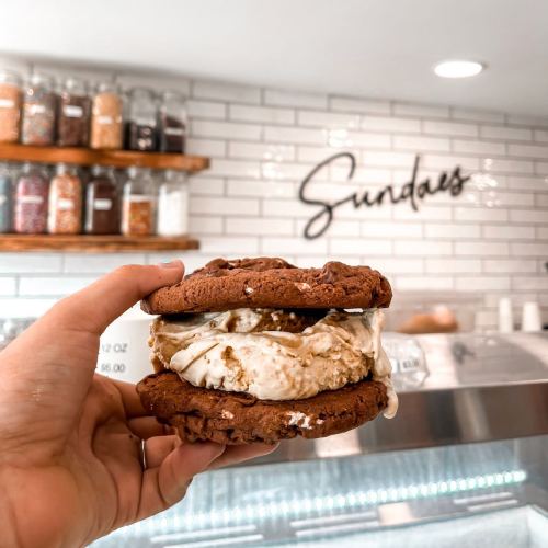 A hand holding a cookie ice cream sandwich in front of a shop with jars on shelves and a sign that says "Sundaes."