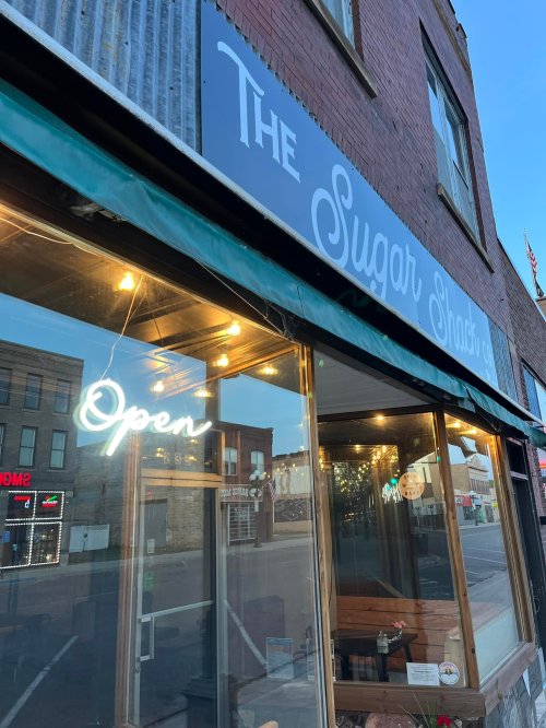 Exterior of The Sugar Shack with a glowing "Open" sign and warm lights, set in a charming downtown area.