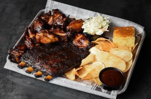A platter featuring ribs, chicken wings, coleslaw, cornbread, chips, and barbecue sauce on a paper-lined tray.