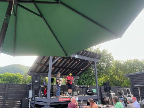A band performs on an outdoor stage under a green umbrella, with an audience seated in front. Trees and hills in the background.