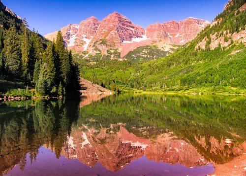 Scenic view of Maroon Bells mountains reflecting in a tranquil lake, surrounded by lush green trees and clear blue sky.