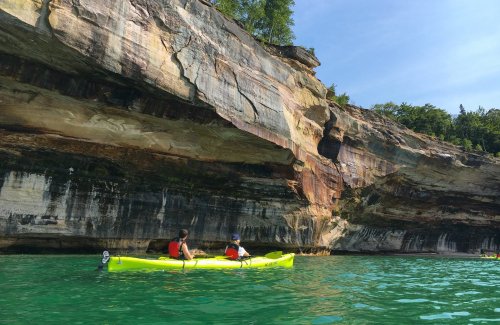 Two people kayak near a rocky cliff with vibrant colors, surrounded by clear turquoise water and lush greenery.
