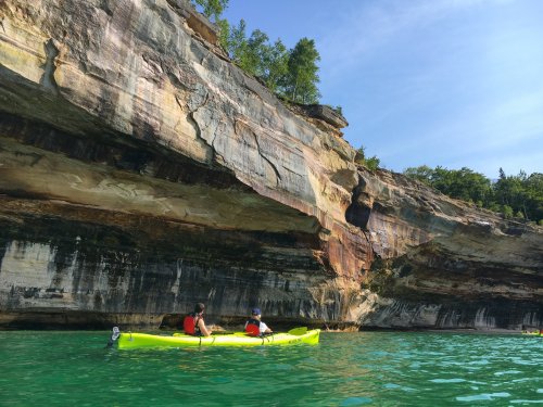 Two kayakers paddle in clear green water near a towering, colorful rock formation under a blue sky.