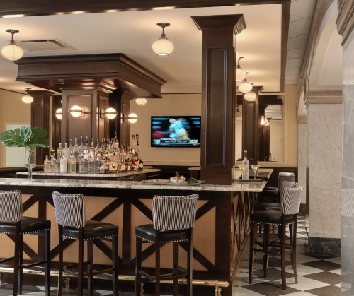 A stylish bar with a marble countertop, wooden accents, and high stools, featuring a TV and a well-stocked liquor shelf.