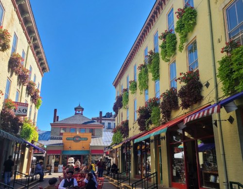 Colorful buildings with hanging flower baskets, bustling market area under a clear blue sky. People enjoying the space.