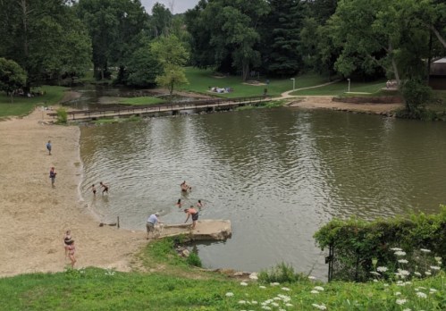 A sandy beach area by a calm lake, with people swimming and relaxing on the shore under green trees.