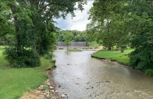 A serene river flows through a lush green landscape, with trees lining the banks and a wooden structure in the background.