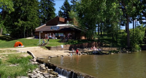 A lakeside scene with a building, sandy area, and people relaxing by the water under sunny skies and trees.