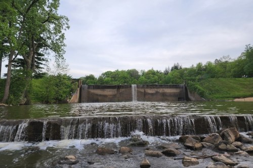 A serene view of a dam with water cascading over it, surrounded by lush greenery and a cloudy sky.
