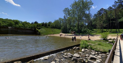 A sunny park scene with a sandy beach, water, trees, and people enjoying the outdoors by a small dam.