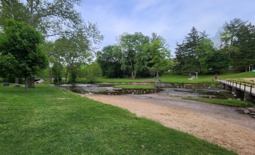A serene park scene featuring a grassy area, a stream, and trees under a clear blue sky.