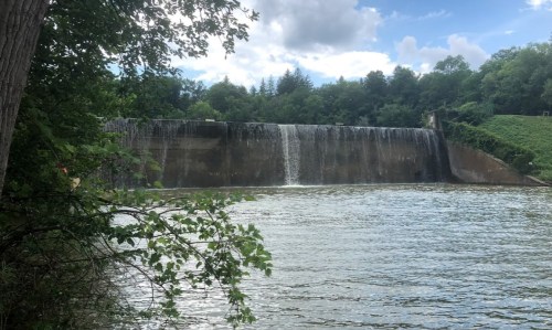 A serene view of a waterfall cascading over a dam, surrounded by lush greenery and a calm river under a blue sky.