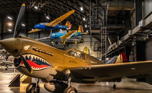 A vintage military aircraft with a shark mouth design in a museum, surrounded by other planes hanging from the ceiling.