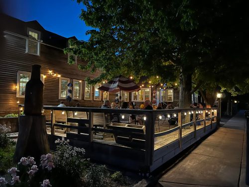 A cozy outdoor dining area with string lights, surrounded by trees, and people enjoying a meal at dusk.