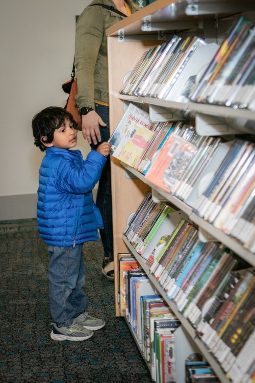 A young child in a blue jacket reaches for a book on a library shelf, with an adult nearby.