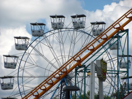 A colorful ferris wheel with white cabins against a cloudy sky, alongside a roller coaster track.