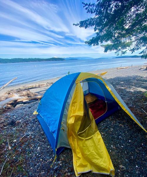 A colorful tent set up on a rocky beach, with a serene view of the water and distant hills under a blue sky.