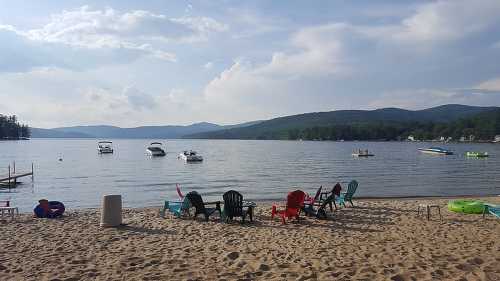 A sandy beach with colorful chairs, boats on the water, and mountains in the background under a partly cloudy sky.